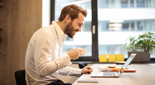 A man sits at an office desk in a well lit room. He smiles as he lift up his coffee cup and reads his laptop screen.